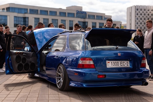 A modified blue sports car with custom wheels and an open door displaying a powerful sound system. Several people stand around, seemingly admiring the vehicle. The car is parked on a paved area in an urban setting with modern buildings in the background.