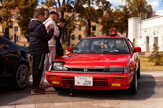 A bright red vintage Honda with a customized license plate reading 'JDM' is parked on a paved area. Three young men are gathered around the car, two of them focusing on their phones. A plush toy sits on the car's roof, and the background features trees and residential buildings.