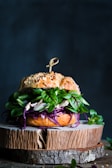 A gourmet sandwich placed on a rustic wooden board, featuring a seeded bun with vibrant green leaves, purple cabbage, and white fillings. The sandwich is garnished with a skewer on top.