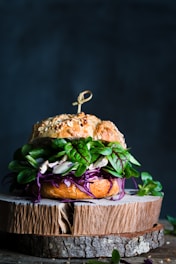 A gourmet sandwich placed on a rustic wooden board, featuring a seeded bun with vibrant green leaves, purple cabbage, and white fillings. The sandwich is garnished with a skewer on top.