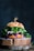 A gourmet sandwich placed on a rustic wooden board, featuring a seeded bun with vibrant green leaves, purple cabbage, and white fillings. The sandwich is garnished with a skewer on top.