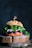 A gourmet sandwich placed on a rustic wooden board, featuring a seeded bun with vibrant green leaves, purple cabbage, and white fillings. The sandwich is garnished with a skewer on top.