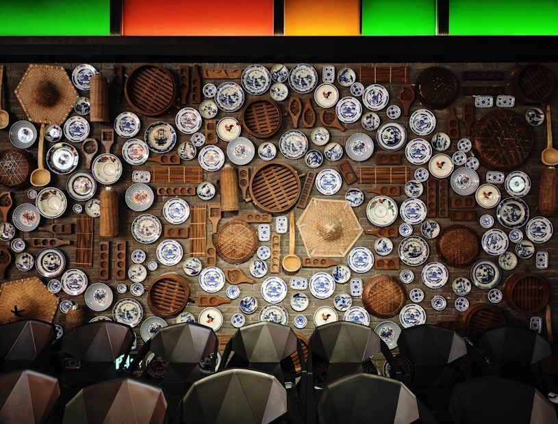 A wall adorned with an eclectic arrangement of decorative blue and white ceramic plates interspersed with wooden kitchen utensils and woven baskets, organized in an artistic manner. The plates feature intricate designs, while the wooden elements add a rustic touch. Overhead, colored panels in green and orange provide a vibrant accent.
