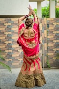 A person stands with their back to the camera, wearing a vibrant, traditional Indian lehenga. The outfit is richly embroidered with golden patterns on a bright pink and deep brown fabric. Their arms are raised gracefully, adorned with colorful bangles and intricately designed henna on the hands. The background features a patterned stone wall and lush greenery.