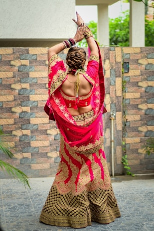 A person stands with their back to the camera, wearing a vibrant, traditional Indian lehenga. The outfit is richly embroidered with golden patterns on a bright pink and deep brown fabric. Their arms are raised gracefully, adorned with colorful bangles and intricately designed henna on the hands. The background features a patterned stone wall and lush greenery.