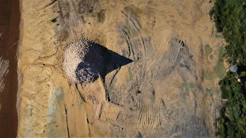 An aerial view of a large mound of gravel or stones positioned in the center of a barren, sandy landscape. The surrounding area features tire tracks and patches of greenery on one side. Shadows indicate the time might be late afternoon.
