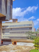A modern, multi-story building with a concrete facade and large windows, labeled 'Union Européenne'. The foreground includes a patio area with scattered colorful seating cubes. The sky is bright blue with scattered clouds, and there is some urban greenery, including trees, around the building.