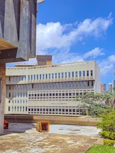 A modern, multi-story building with a concrete facade and large windows, labeled 'Union Européenne'. The foreground includes a patio area with scattered colorful seating cubes. The sky is bright blue with scattered clouds, and there is some urban greenery, including trees, around the building.