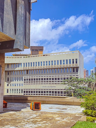 A modern, multi-story building with a concrete facade and large windows, labeled 'Union Europ&eacute;enne'. The foreground includes a patio area with scattered colorful seating cubes. The sky is bright blue with scattered clouds, and there is some urban greenery, including trees, around the building.