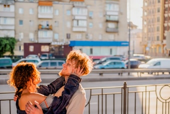 A couple is sharing a joyful and intimate moment outdoors on a sunny day. They are positioned near a street with parked cars and buildings in the background. The lighting creates a warm atmosphere, highlighting their expressions and capturing the lively energy of the scene.