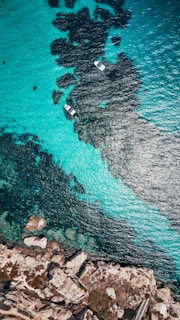 An aerial view of turquoise ocean waters with visible dark patches beneath the surface, likely indicating rocks or seaweed. Two small boats can be seen floating amidst the clear waters. On one side, rugged coastline with rocky formations and sparse vegetation is visible.