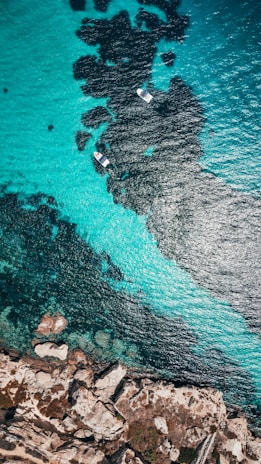 An aerial view of turquoise ocean waters with visible dark patches beneath the surface, likely indicating rocks or seaweed. Two small boats can be seen floating amidst the clear waters. On one side, rugged coastline with rocky formations and sparse vegetation is visible.