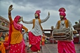 Statues of people in traditional Punjabi attire, engaged in a dance with vibrant clothing in yellow, white, and pink colors. The male figures wear turbans and are playing musical instruments, including a dhol. The scene appears lively and culturally rich, set against a backdrop of a building with rustic charm, under a cloudy sky.
