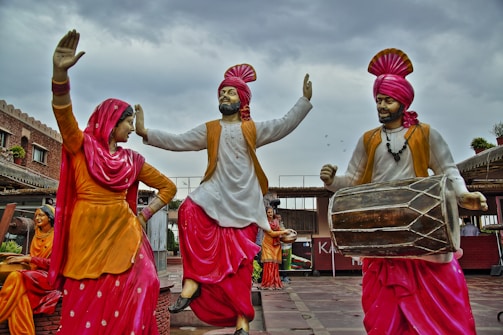 Statues of people in traditional Punjabi attire, engaged in a dance with vibrant clothing in yellow, white, and pink colors. The male figures wear turbans and are playing musical instruments, including a dhol. The scene appears lively and culturally rich, set against a backdrop of a building with rustic charm, under a cloudy sky.