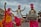 Statues of people in traditional Punjabi attire, engaged in a dance with vibrant clothing in yellow, white, and pink colors. The male figures wear turbans and are playing musical instruments, including a dhol. The scene appears lively and culturally rich, set against a backdrop of a building with rustic charm, under a cloudy sky.