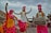Statues of people in traditional Punjabi attire, engaged in a dance with vibrant clothing in yellow, white, and pink colors. The male figures wear turbans and are playing musical instruments, including a dhol. The scene appears lively and culturally rich, set against a backdrop of a building with rustic charm, under a cloudy sky.