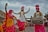 Statues of people in traditional Punjabi attire, engaged in a dance with vibrant clothing in yellow, white, and pink colors. The male figures wear turbans and are playing musical instruments, including a dhol. The scene appears lively and culturally rich, set against a backdrop of a building with rustic charm, under a cloudy sky.