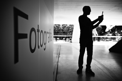 A silhouette of a person holding a phone stands in a spacious, modern interior with large windows allowing natural light. On the left, a wall displays the word 'Fotografia'. The atmosphere is serene with a focus on photography.