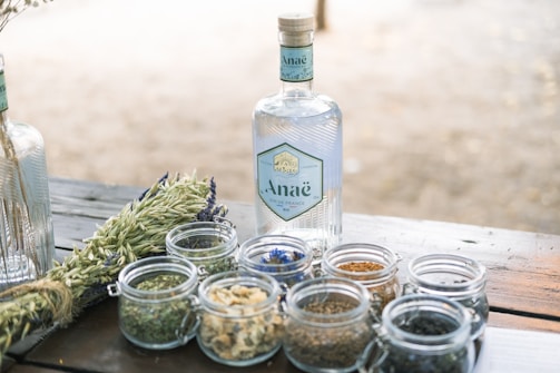 A bottle of gin with a label reading 'Anaë' is placed on a wooden table alongside small glass jars containing various herbs and spices. The background is softly blurred, suggesting an outdoor setting. Fresh green plants are also visible next to the jars, contributing to a natural and organic vibe.