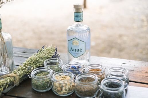 A bottle of gin with a label reading 'Ana&euml;' is placed on a wooden table alongside small glass jars containing various herbs and spices. The background is softly blurred, suggesting an outdoor setting. Fresh green plants are also visible next to the jars, contributing to a natural and organic vibe.