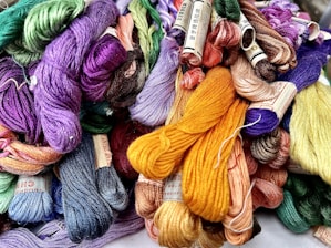 A collection of colorful embroidery threads is bundled together, featuring a wide array of hues including purples, greens, yellows, and blues. Each skein is neatly wound and many have labels attached.