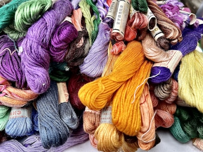 A collection of colorful embroidery threads is bundled together, featuring a wide array of hues including purples, greens, yellows, and blues. Each skein is neatly wound and many have labels attached.