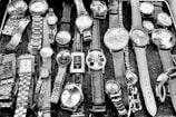 A collection of various wristwatches displayed on a surface. The watches vary in style, size, and design, featuring leather, metal, and chain straps. Some have round faces, while others have square ones with different clock face designs, including numerals and abstract patterns.