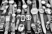 A collection of various wristwatches displayed on a surface. The watches vary in style, size, and design, featuring leather, metal, and chain straps. Some have round faces, while others have square ones with different clock face designs, including numerals and abstract patterns.