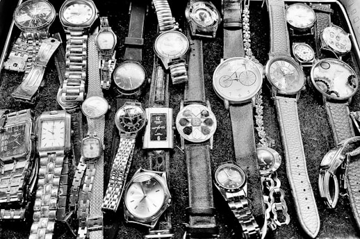 A collection of various wristwatches displayed on a surface. The watches vary in style, size, and design, featuring leather, metal, and chain straps. Some have round faces, while others have square ones with different clock face designs, including numerals and abstract patterns.