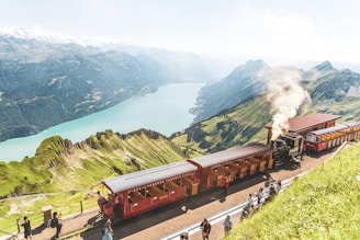 A steam train travels along a hillside railway offering a stunning view of a turquoise lake surrounded by majestic mountains. Passengers are visible on the platform, and vibrant green grass covers the landscape.