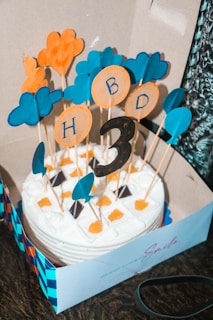 A round cake in a box is decorated with white frosting and small orange and chocolate toppings. Several colorful paper cutouts, including clouds and circles with letters, are attached to sticks, protruding from the cake. The letters 'HBD' and the number '3' are visible among the decorations, indicating a third birthday celebration. The box has a blue and orange checkered pattern on the side.