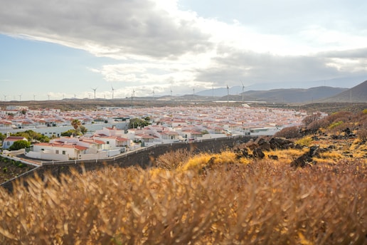 A landscape featuring a residential area with white houses and terracotta roofs surrounded by dry grassland and rocky terrain. In the background, wind turbines are visible against a partly cloudy sky, while distant hills rise on the horizon.