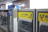 Glass safety doors at a train platform with prominent yellow warning signs indicating to mind the platform gap. The signs have both text and pictograms, and another sign designates the area as Car 6.