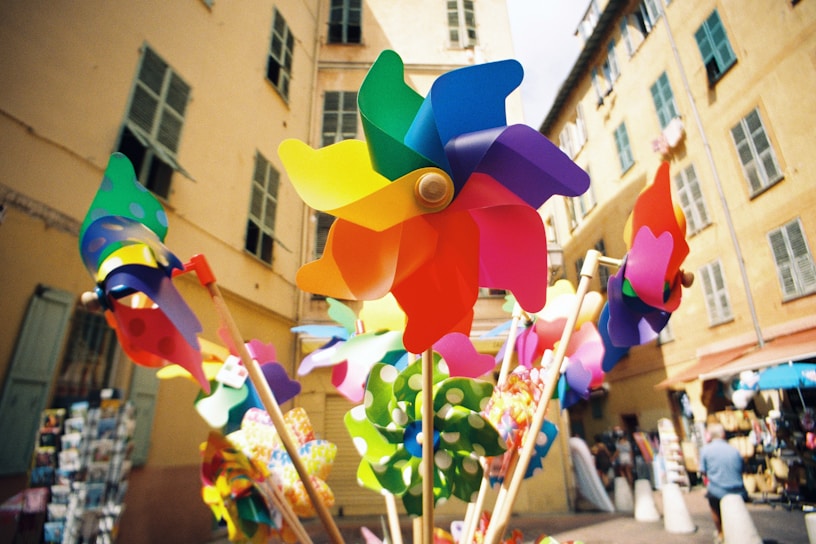 Colorful pinwheels are on display, set against the backdrop of a sunlit urban street with tall, muted buildings. The pinwheels are vibrant and varied in shape, adding a whimsical and lively touch to the scene.