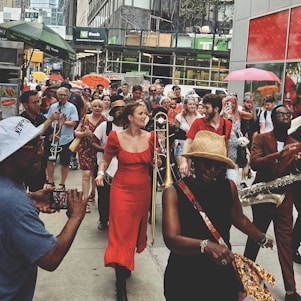 A lively street parade features musicians playing instruments like trombone and saxophone. People in vibrant attire, including a woman in a red dress and a man in a brown suit, move through a bustling city sidewalk. Some carry umbrellas, while onlookers capture the moment with phones. The atmosphere is festive, with a diverse crowd of participants and spectators enjoying the event.