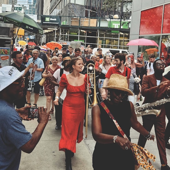 A lively street parade features musicians playing instruments like trombone and saxophone. People in vibrant attire, including a woman in a red dress and a man in a brown suit, move through a bustling city sidewalk. Some carry umbrellas, while onlookers capture the moment with phones. The atmosphere is festive, with a diverse crowd of participants and spectators enjoying the event.