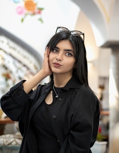 A woman with long dark hair and dark eyes is wearing a black outfit and glasses on top of her head, posing with one hand resting on her face. She is standing in a well-lit indoor space with a curved staircase in the background and colorful floral decorations on the wall.