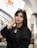 A woman with long dark hair and dark eyes is wearing a black outfit and glasses on top of her head, posing with one hand resting on her face. She is standing in a well-lit indoor space with a curved staircase in the background and colorful floral decorations on the wall.