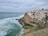A coastal scene featuring a collection of whitewashed buildings with red-tiled roofs situated on a cliffside. The ocean waves crash against the rocky shore, while a small, natural-looking swimming pool faces the sea. The sky is overcast, giving a calm yet slightly dramatic atmosphere.