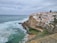 A coastal scene featuring a collection of whitewashed buildings with red-tiled roofs situated on a cliffside. The ocean waves crash against the rocky shore, while a small, natural-looking swimming pool faces the sea. The sky is overcast, giving a calm yet slightly dramatic atmosphere.