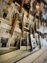 A collection of Polaroid photographs is clipped onto a string with wooden clothespins. The photos appear to be portraits, some with a mugshot-style backdrop, including one with a cartoon face and several showing people holding signs.