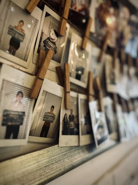A collection of Polaroid photographs is clipped onto a string with wooden clothespins. The photos appear to be portraits, some with a mugshot-style backdrop, including one with a cartoon face and several showing people holding signs.