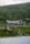 A large cabin or lodge is nestled in a lush, mountainous area, surrounded by dense green forest and rocky terrain. The building features a dark, rustic architecture with a gabled roof. In the foreground, a calm body of water reflects the serene landscape.