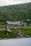 A large cabin or lodge is nestled in a lush, mountainous area, surrounded by dense green forest and rocky terrain. The building features a dark, rustic architecture with a gabled roof. In the foreground, a calm body of water reflects the serene landscape.
