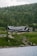 A large cabin or lodge is nestled in a lush, mountainous area, surrounded by dense green forest and rocky terrain. The building features a dark, rustic architecture with a gabled roof. In the foreground, a calm body of water reflects the serene landscape.
