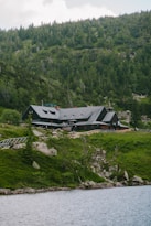 A large cabin or lodge is nestled in a lush, mountainous area, surrounded by dense green forest and rocky terrain. The building features a dark, rustic architecture with a gabled roof. In the foreground, a calm body of water reflects the serene landscape.