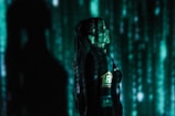 A person stands in profile against a backdrop of cascading green digital code, reminiscent of a futuristic or cyberpunk aesthetic. The person is wearing dark clothing with visible logos and has long hair. Shadows and green light interplay across their face and body, creating a mysterious atmosphere.