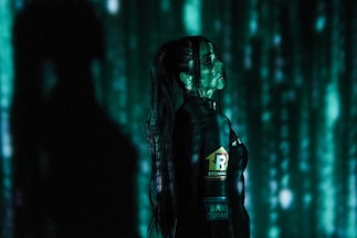 A person stands in profile against a backdrop of cascading green digital code, reminiscent of a futuristic or cyberpunk aesthetic. The person is wearing dark clothing with visible logos and has long hair. Shadows and green light interplay across their face and body, creating a mysterious atmosphere.