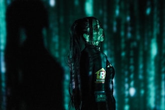 A person stands in profile against a backdrop of cascading green digital code, reminiscent of a futuristic or cyberpunk aesthetic. The person is wearing dark clothing with visible logos and has long hair. Shadows and green light interplay across their face and body, creating a mysterious atmosphere.