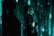 A person stands in profile against a backdrop of cascading green digital code, reminiscent of a futuristic or cyberpunk aesthetic. The person is wearing dark clothing with visible logos and has long hair. Shadows and green light interplay across their face and body, creating a mysterious atmosphere.